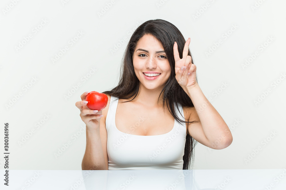 Young hispanic woman holding a tomato showing victory sign and smiling broadly. Young hispanic woman holding a tomato showing victory sign and smiling broadly.