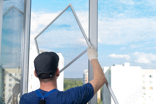 a worker in a blue uniform, rear view, installs a mosquito net in a plastic window frame, against the sky