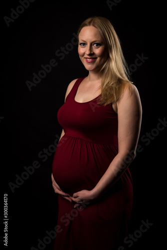 Pregnant woman in red dress holding belly on black background.