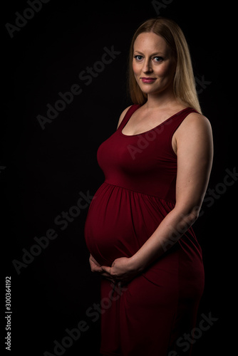 Pregnant woman in red dress holding belly on black background.