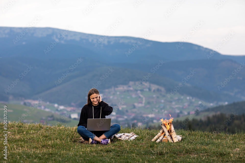 Young woman with laptop in the mountains. Girl works while sitting on the grass, the bonfire is lit on the side. Work, business, freelance. Place for inscription. Young woman with laptop in the mountains. Girl works while sitting on the grass, the bonfire is lit on the side. Work, business, freelance. Place for inscription.
