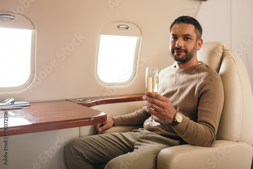 cheerful man holding champagne glass while sitting in first class of private jet