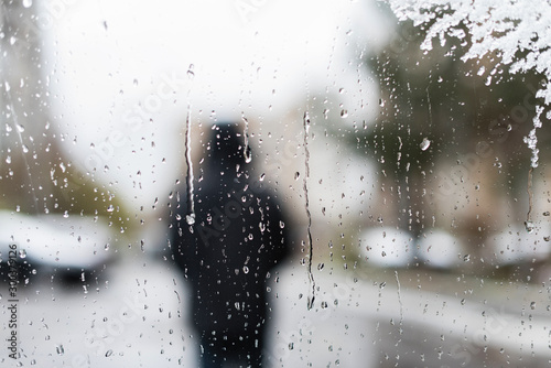 Unfocused silhouette man in a winter jacket with a hood behind car window with melted snow texture. Winter background