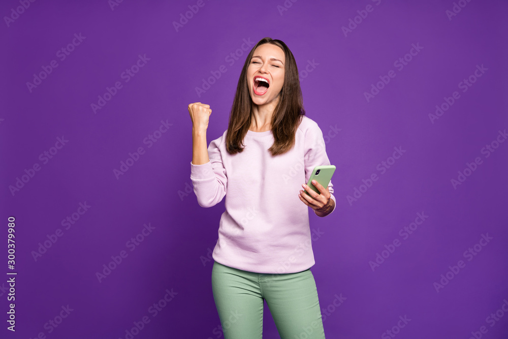 Photo of excited crazy cheerful ecstatic overjoyed girlfriend expressing emotions on face making fist holding phone screaming with rejoicing feeling isolated over purple vivid color background