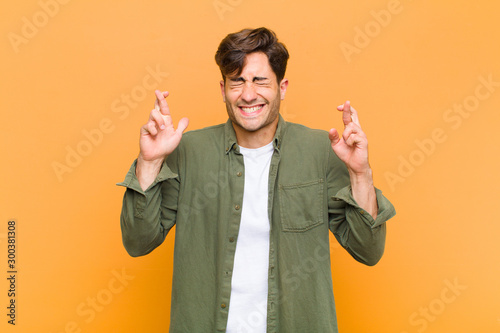 young handsome man smiling and anxiously crossing both fingers, feeling worried and wishing or hoping for good luck against orange background photo