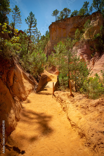 View over the ochre pathway and orange rocks, yellow sand and green pinetrees with a wooden bridge against a blue sky in Roussillion, Vaucluse-Luberon, France in the summer.