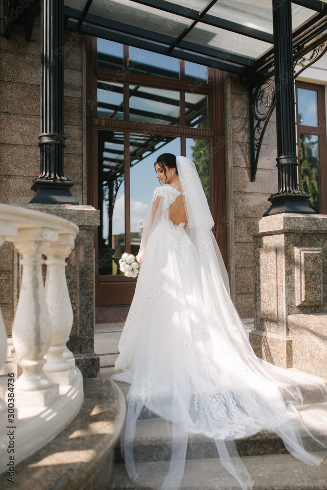 Obraz premium Back view of Gorgeous bride stand on the stairs in elegant wedding dress with bouquet of white flowers in her wedding day