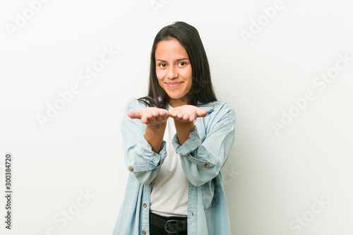 Young hispanic woman holding something with palms, offering to camera.