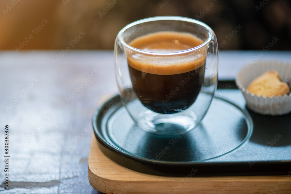 Hot espresso coffee in a clear glass, placed on a metal tray and wooden layer beside the coffee cup with a bear shaped cookie . All laid on dark brown wooden tables in retro coffee shop.