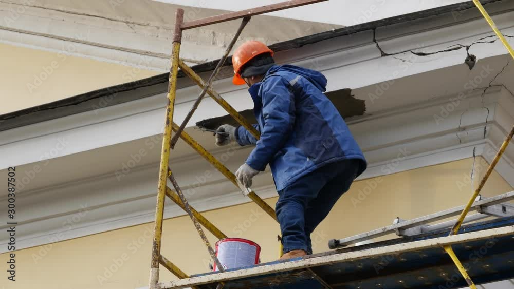 A construction worker on a scaffolding plaster decor on a building. Daytime. Bucket with putty. Protective orange helmet on the head