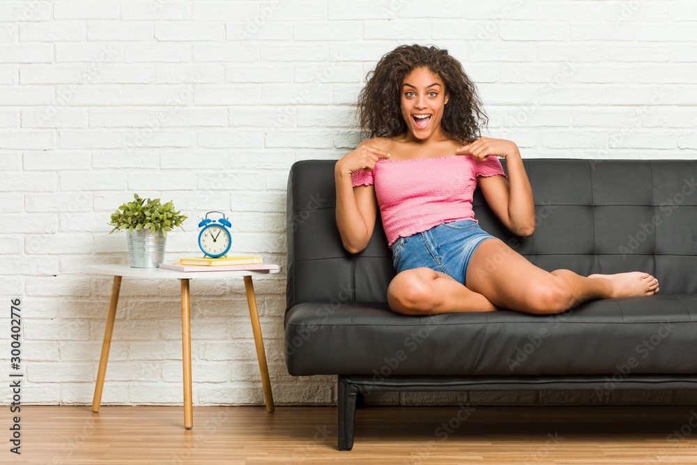 © Asier - Young african american woman sitting on the sofa surprised pointing with finger, smiling broadly. © Asier - Young african american woman sitting on the sofa surprised pointing with finger, smiling broadly.