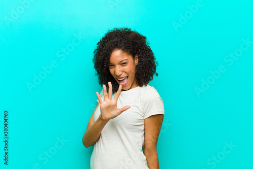 young black woman smiling and looking friendly, showing number five or fifth with hand forward, counting down against blue wall photo