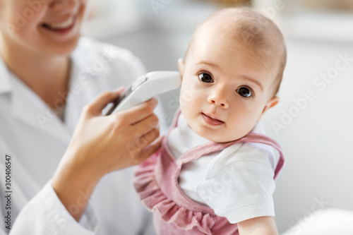 medicine  healthcare and pediatrics concept - female pediatrician doctor with digital infrared thermometer measuring baby girl patient s ear temperature at clinic or hospital