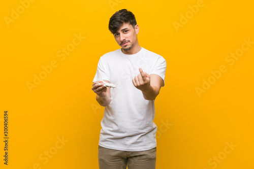 Young hispanic man holding an airplane icon pointing with finger at you as if inviting come closer.