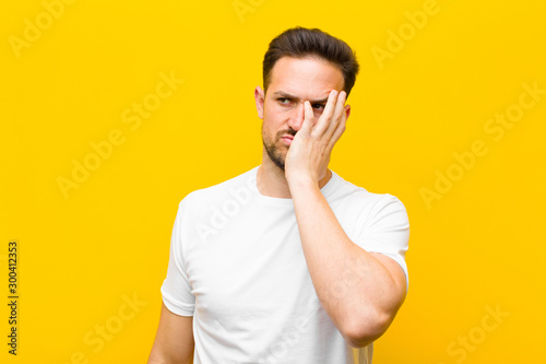 young handsome man feeling bored, frustrated and sleepy after a tiresome, dull and tedious task, holding face with hand against orange wall photo