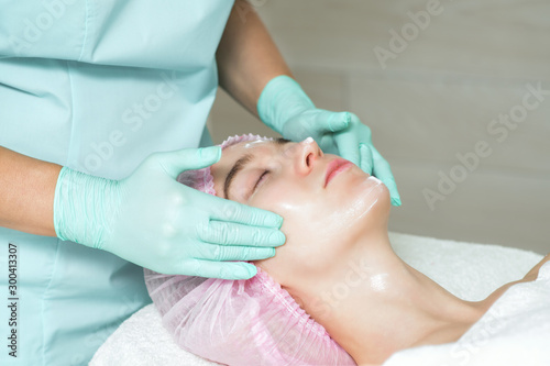 Hands of beautician is applying a white cream to the woman face in the spa beauty salon.