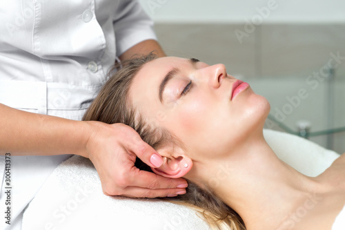 Young caucasian woman lying down receiving a head massage by female beautician touching woman s ears and temples in the spa beauty salon close up.