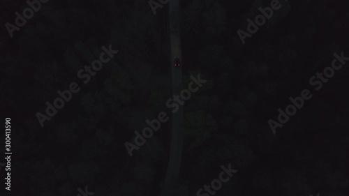 Aerial front view black car driving on dark countryside asphalt road among summer forest at night. View from drone car with headlights riding on night road through dark trees