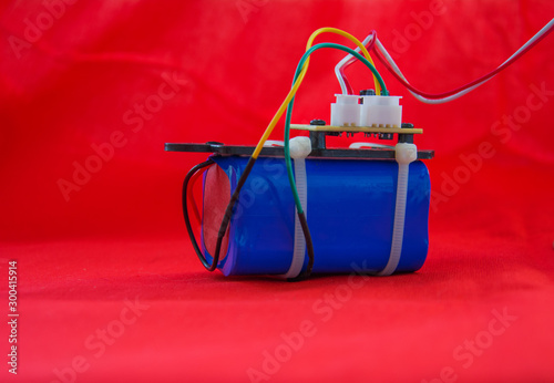 Part of an electronic device. Blue battery pack with printed circuit Board fastened by zip tie, connectors and colored wires on a red background.