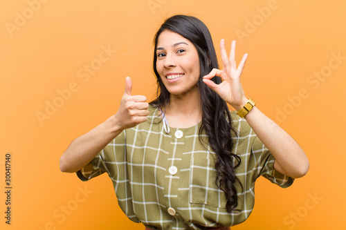 young pretty hispanic woman feeling happy, amazed, satisfied and surprised, showing okay and thumbs up gestures, smiling against brown wall photo