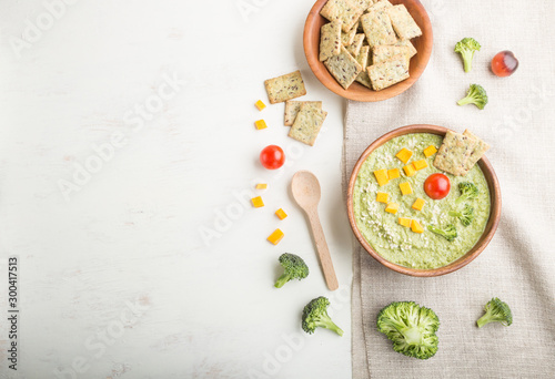 Green broccoli cream soup with crackers and cheese in wooden bowl on a white wooden background. top view, copy space.