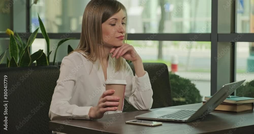 Businesswoman holds in her hand plastic coffee cup, using laptop pc indoors in coffee house. Young adult working online. Female smiling at camera Businesswoman holds in her hand plastic coffee cup, using laptop pc indoors in coffee house. Young adult working online. Female smiling at camera