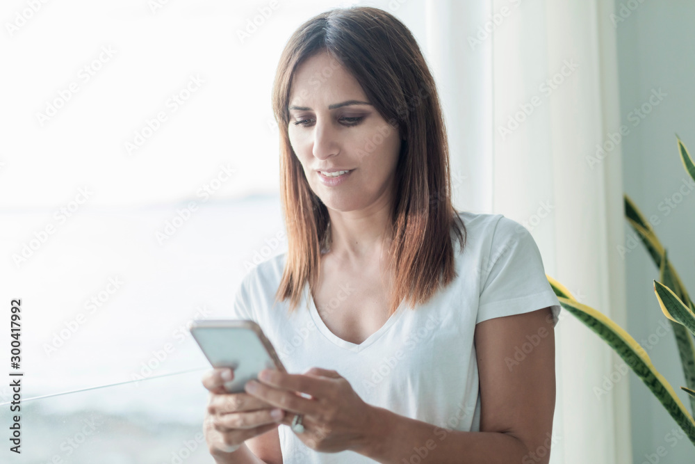 Happy adult woman using a mobile phone while standing next to a window Happy adult woman using a mobile phone while standing next to a window