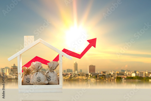 Money bags in the wooden home model and red arrow graph with growing value on photo blur cityscape background, Loan and business investment for real estate concept.