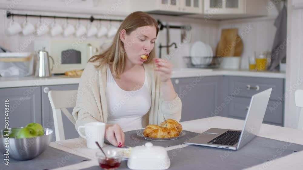 Tracking shot of woman sitting at table and using laptop while eating buttered croissant and drinking coffee Tracking shot of woman sitting at table and using laptop while eating buttered croissant and drinking coffee