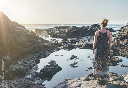 blonde woman in summer dress standing at rocky shore looking at ocean in sunlight