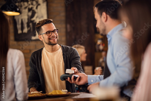 Obraz na płótnie Young smiling man using mobile phone while paying a bill in a restaurant
