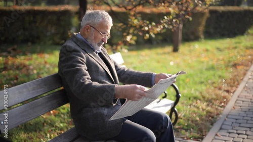 Handsome grandfather with a beautiful beard in a gray jacket sits on a bench in the park and reads a newspaper. Senior gray-haired man in glasses
