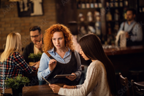 Redhead waitress talking to a woman while taking an order in a pub.