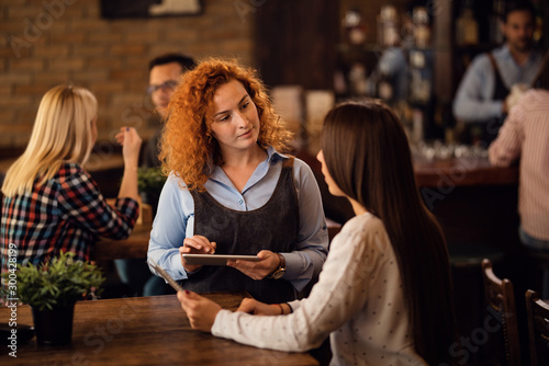 Young waitress taking order on touchpad while communicating with female customer.