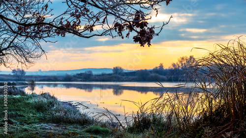 Scenic landscape with river, trees and frost on grass during sunrise in autumn_