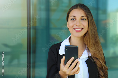 Happy young business woman checking her smart phone and looking at camera out of office. Copy space. Free Download Happy young business woman checking her smart phone and looking at camera out of office. Copy space. - GFXLoad