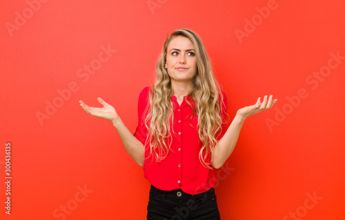 young blonde woman feeling puzzled and confused, doubting, weighting or choosing different options with funny expression against red wall