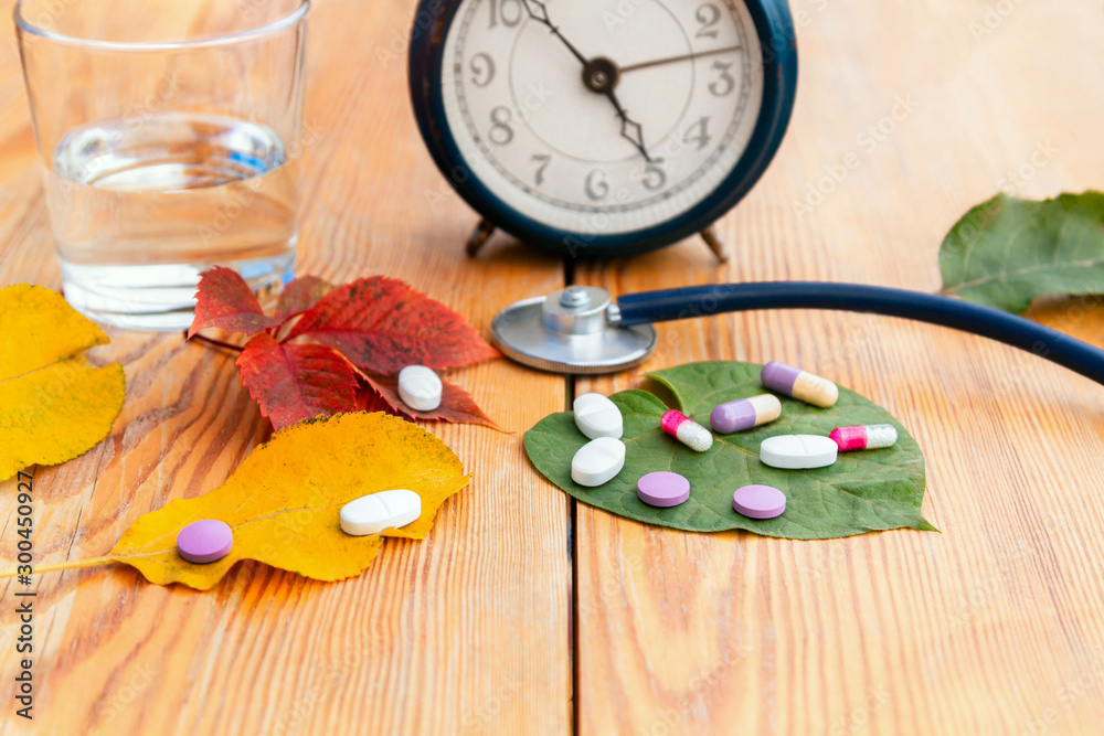 Autumn colds. Time to take pills. Alarm clock, stethoscope, colorful pills, autumn leaves of different colors on a wooden table. Autumn colds. Time to take pills. Alarm clock, stethoscope, colorful pills, autumn leaves of different colors on a wooden table.
