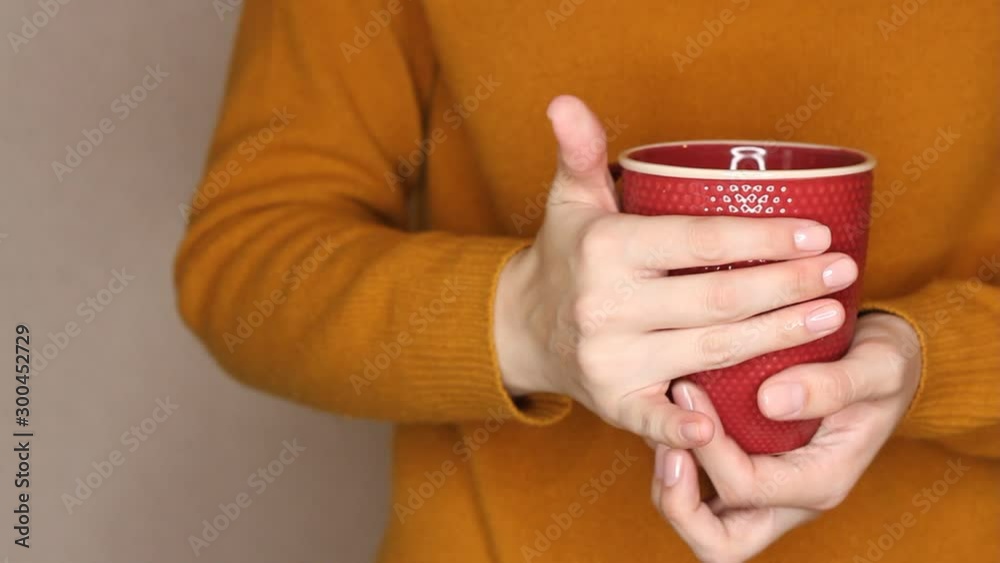 Young woman in yellow sweater is holding red cup of tea or coffee. Changing hand, trying to warm hands with hot drink Young woman in yellow sweater is holding red cup of tea or coffee. Changing hand, trying to warm hands with hot drink