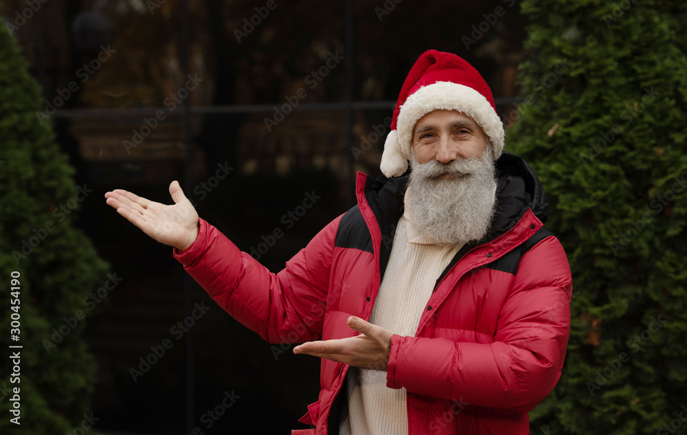 Obraz premium Senior man with beard in red santa hat presenting something outdoor. Xmas. New Year.