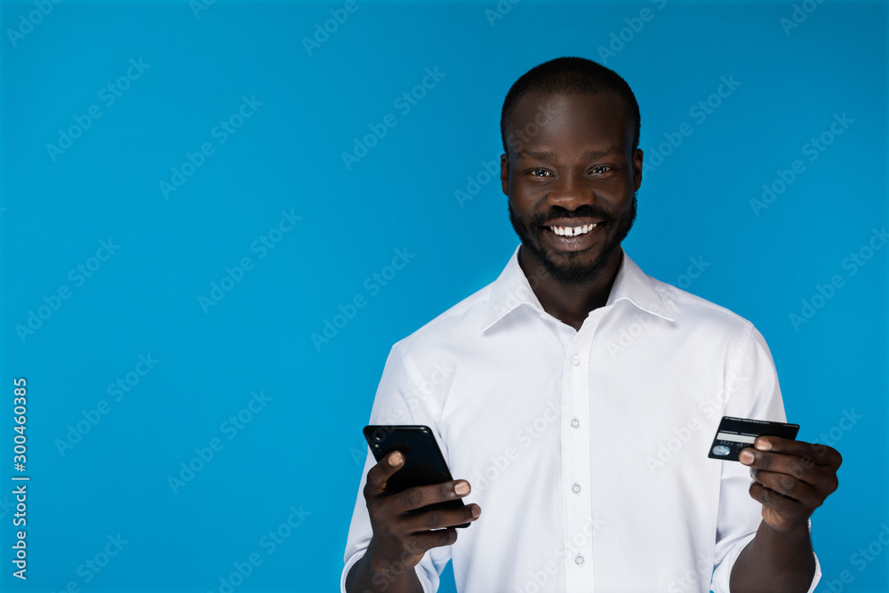 Smiling american black man looks at the camera and holds a phone and credit card
