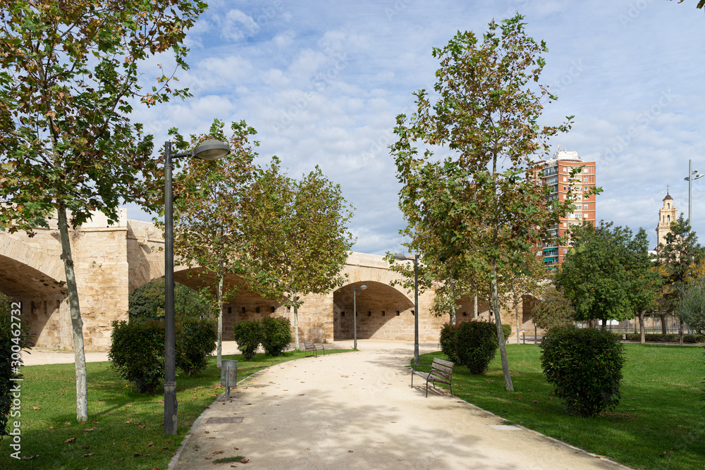 Path with bench towards ancient stone bridge across old riverbed. Puente del Real, Turia river, Valencia, Spain Path with bench towards ancient stone bridge across old riverbed. Puente del Real, Turia river, Valencia, Spain