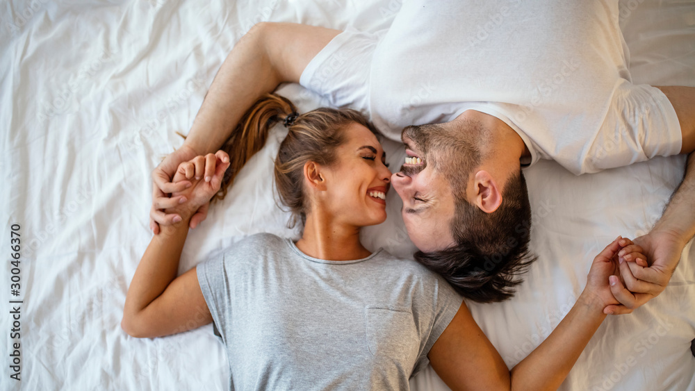 Guy and a girl in a cozy home environment. Happy man and woman lying in the bedroom stock photo. Top view of smiling young couple cuddling in bed in morning.  Beautiful pair of lovers hug and kiss - obrazy, fototapety, plakaty 