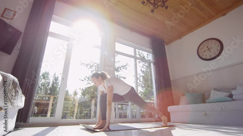 Tracking shot with low angle of woman in leggings and t-shirt doing yoga poses in living room on sunny morning photo