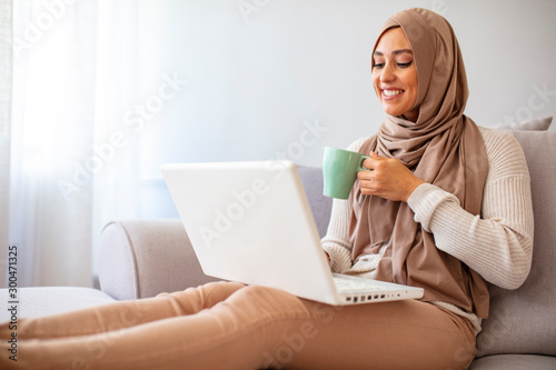 Happy muslim woman wearing hijab at home. Girl drinking coffee and shopping online on laptop, copy space. Shot of a young businesswoman using a laptop and having coffee at home.