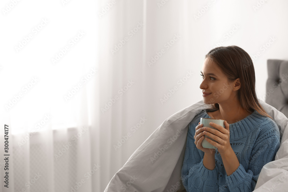 Young woman in warm sweater with hot drink on bed at home. Space for text Young woman in warm sweater with hot drink on bed at home. Space for text