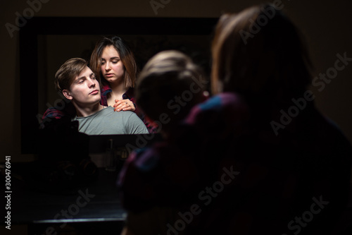 Reflection of a man and a girl in the mirror. The girl puts her arm around the man's shoulders and looks closely at him. People on a dark background. The tension in the relationship. Incomprehension.