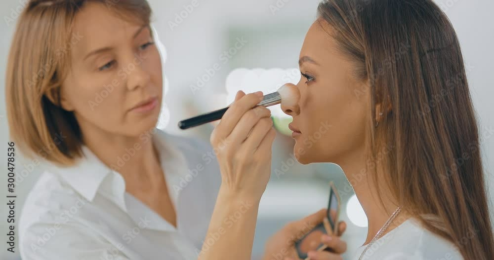 Professional makeup artist applies powder, face makeup with a brush. Beautiful girl sitting in front of a large mirror in a beauty salon. Portrait view. Professional makeup artist applies powder, face makeup with a brush. Beautiful girl sitting in front of a large mirror in a beauty salon. Portrait view.