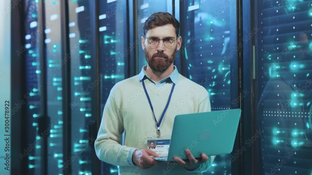 Bearded smart IT specialist working on laptop standing at operational rack server cabinets in data center. Computer service. Telecommunication concept. Bearded smart IT specialist working on laptop standing at operational rack server cabinets in data center. Computer service. Telecommunication concept.