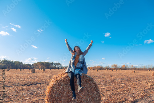 A girl of European appearance in a gray coat stands in a field near a larger bale with hay against a blue sky.